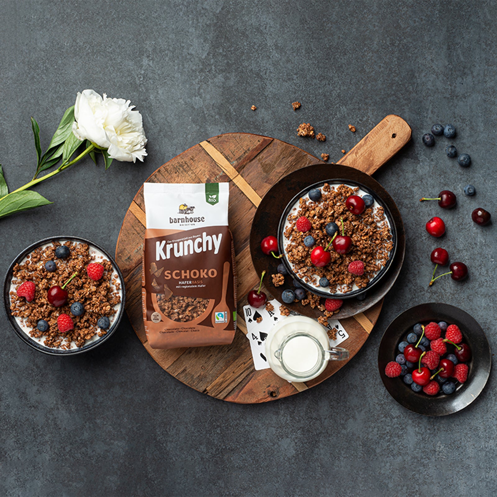 Blick von oben auf eine runde Holzplatte mit Pokerkarten & einer Karaffe Milch. Auch ein braun-weißer Beutel Krunchy Schoko von Knuspermüsli-Hersteller Barnhouse liegt mit drauf, ebenso wie eine Müslischale mit dem Schoko-Müsli, Milch & Topping aus Kirschen, Himbeeren & Heidelbeeren. Von links ragt eine weiße Blume mit grünem Stiel herein. Links ist eine weitere Schale mit gleichem Inhalt. Rechts liegen Kirschen, Himbeeren und Heidelbeeren auf der schwarzen Marmorplatte verteilt & in einer schwarzen Schale.