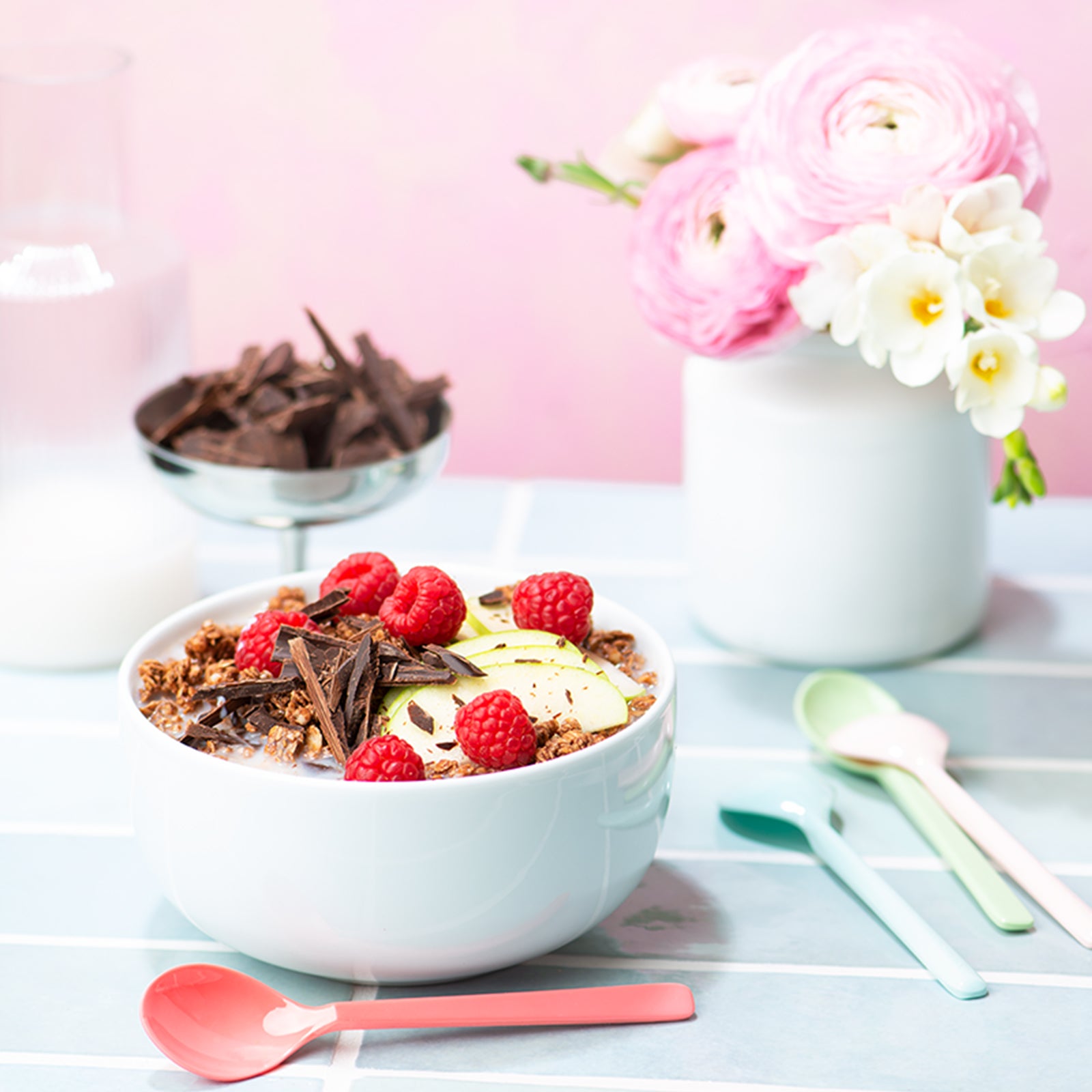 Blick von vorne auf einen Frühstückstisch mit einer weißen Müslischale, die mit Milch und Krunchy Schoko von Bio-Knuspermüsli-Hersteller Barnhouse befüllt ist. Als dekoratives Highlight liegen noch Schokosplitter, Himbeeren und grüne Apfelscheiben mit drauf. Auf dem gefliesten Tisch steht eine silberne Schale mit Schokosplittern sowie eine weiße Vase mit rosa und weißen Blumen. Vorne liegen noch vier Löffel in verschiedenen Farben.