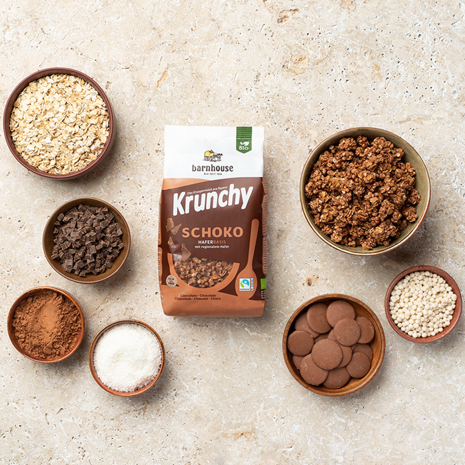 Blick von oben auf einen braun-weißen Beutel Krunchy Schoko von Bio-Knuspermüsli-Hersteller Barnhouse auf heller Marmorplatte. Außenrum stehen sechs braune Schalen mit Haferflocken, Schoko-Stückchen, Kakaopulver, Kokosraspeln, Schokoplättchen und Reispops. Eine weitere Schale ist mit dem Schoko-Knuspermüsli befüllt.