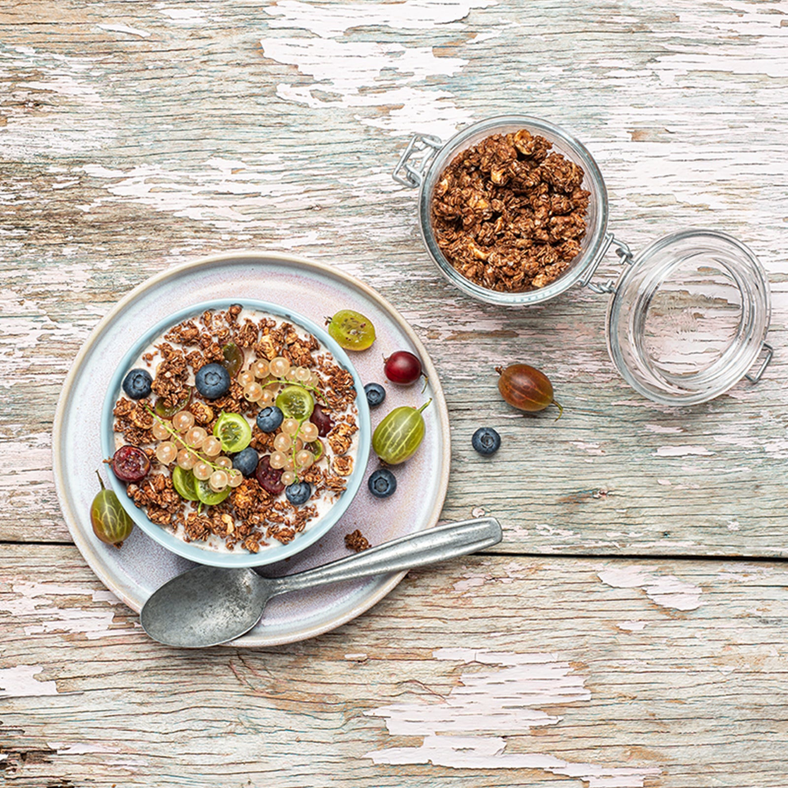 Blick von oben auf einen weiß gestrichenen Holztisch, der schon etwas abblättert. Oben steht ein geöffnetes Vorratsglas mit Krunchy Schoko-Nuss von Bio-Knuspermüsli-Hersteller Barnhouse. Darunter liegen ein Weintraube und eine Heidelbeere. Der Fokus liegt auf der Bowl, die mit dem nussigen Schokoaden-Müsli und Milch befüllt ist. Als Topping liegen Heidelbeeren, weiße Johannisbeeren sowie grüne und rote Stachelbeeren obendrauf. Auf dem Unterteller liegen noch weitere Beeren sowie ein silberner Löffel.