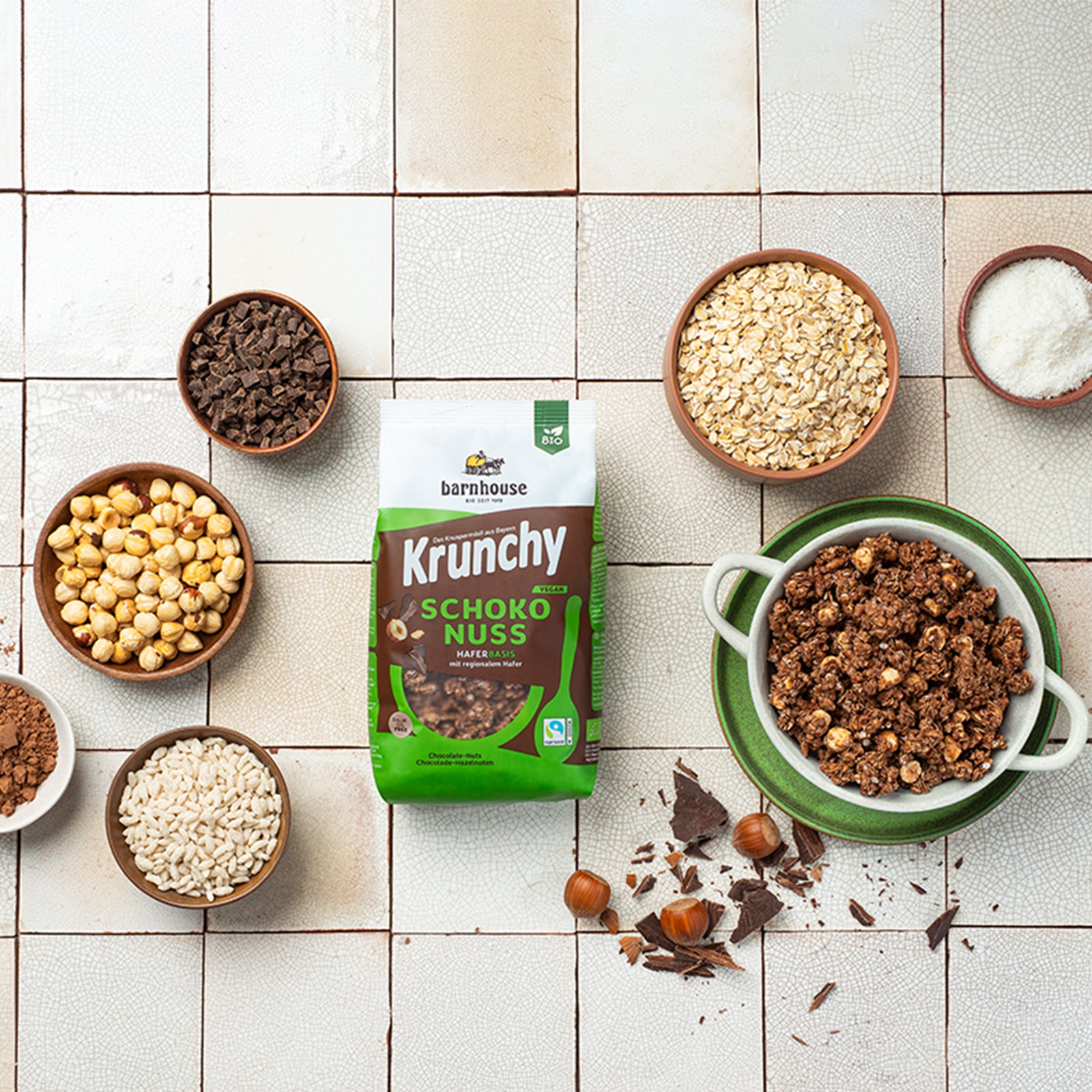 Blick von oben auf einen grün-braun-weißen Beutel Krunchy Schoko-Nuss von Bio-Knuspermüsli-Hersteller Barnhouse auf hellen, gemusterten Fliesen. Außenrum stehen sechs braune Schalen mit Kakaopulver, Reispops, Haselnüsse, Zartbitterschokolade, Kokosraspeln und Haferflocken. Rechts steht auf einem grünen Unterteller eine weiße Suppenschüssel, die mit dem nussigen Schoko-Knuspermüsli befüllt ist. Daneben liegen ein paar Haselnüsse und Schokosplitter.
