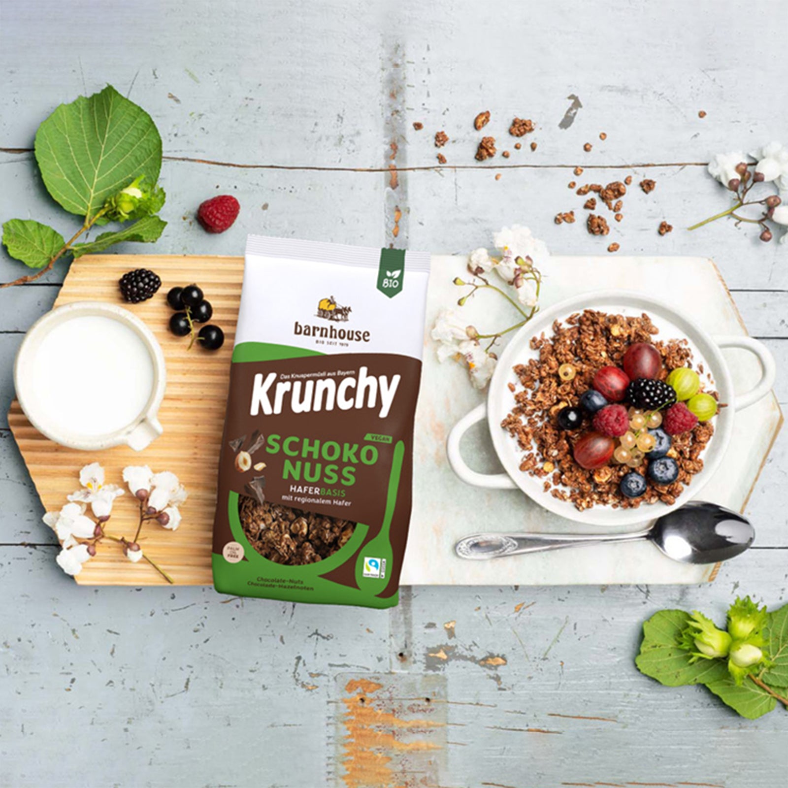 Holzbrett, das auf einem blau angestrichenen Holztisch liegt, der schon etwa abblättert. In der Mitte liegt ein Beutel Krunchy Schoko-Nuss von Bio-Knuspermüsli-Hersteller Barnhouse, daneben eine Kanne Milch und ein silberner Löffel. Außerdem ein paar Blüten, Brom-& Johannisbeeren & ein Suppenteller, der mit Milch & Krunchy Schoko-Nuss befüllt ist - mit Topping aus Weintrauben, Himbeeren, Brombeeren, Heidelbeeren & Johannisbeeren. Auf dem Tisch liegen noch grüne Blätter, eine Himbeere und Müsli-Cluster.