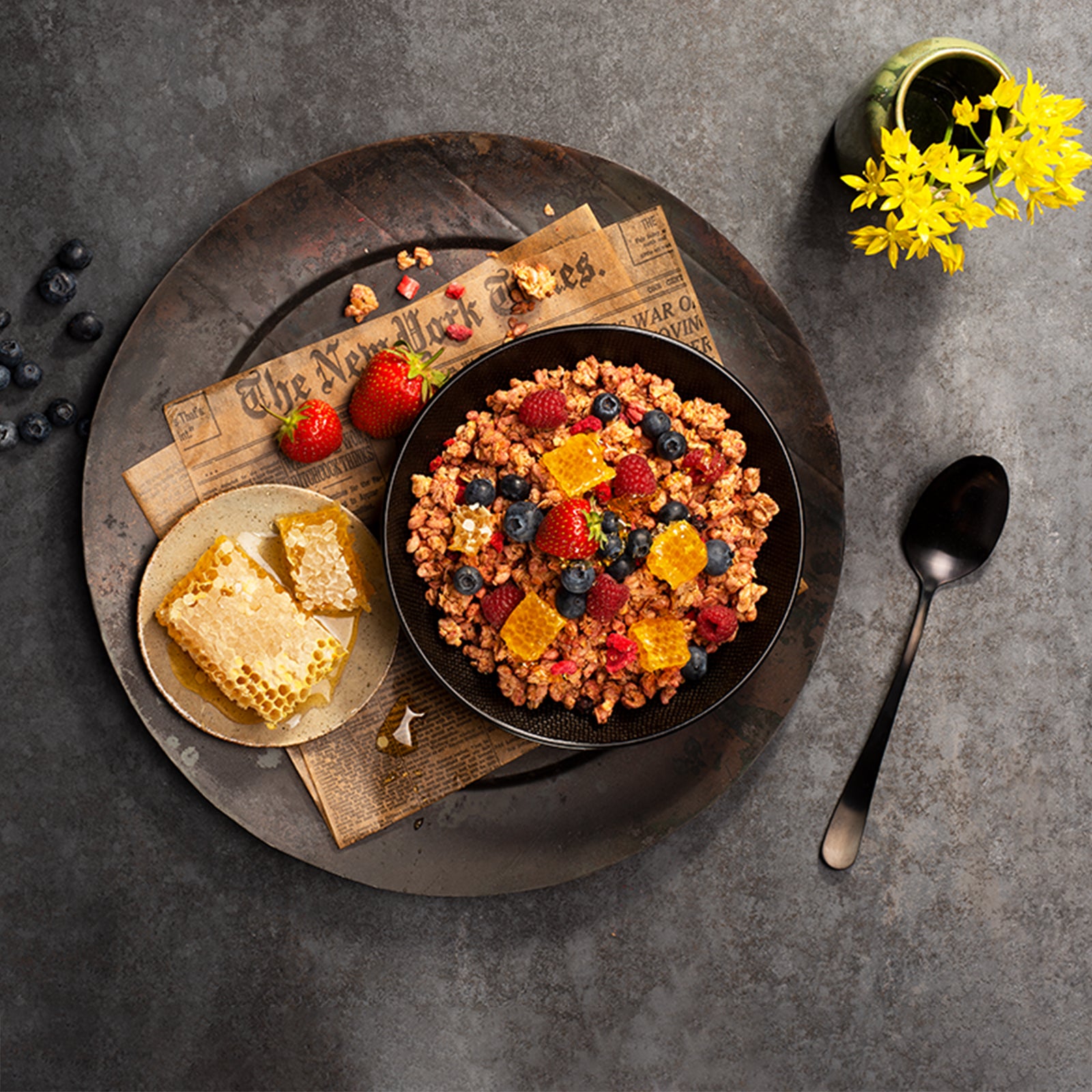 Blick von oben auf eine schwarze Marmorplatte. Links ein paar Heidelbeeren, rechts ein schwarzer Löffel sowie eine grüne Vase mit gelben Blüten. In der Mitte ein runder, dunkler Teller mit alter New York Times Zeitung. Darauf liegen paar Erdbeeren und Müsli-Cluster sowie ein weißer Teller mit zwei Honigwarben. Der Fokus liegt auf einem schwarzen Teller, der mit Krunchy Waldbeere alternativ gesüßt von Bio-Knuspermüsli-Hersteller Barnhouse befüllt & mit Waldbeeren sowie Honigwarben aufgewertet wurde.
