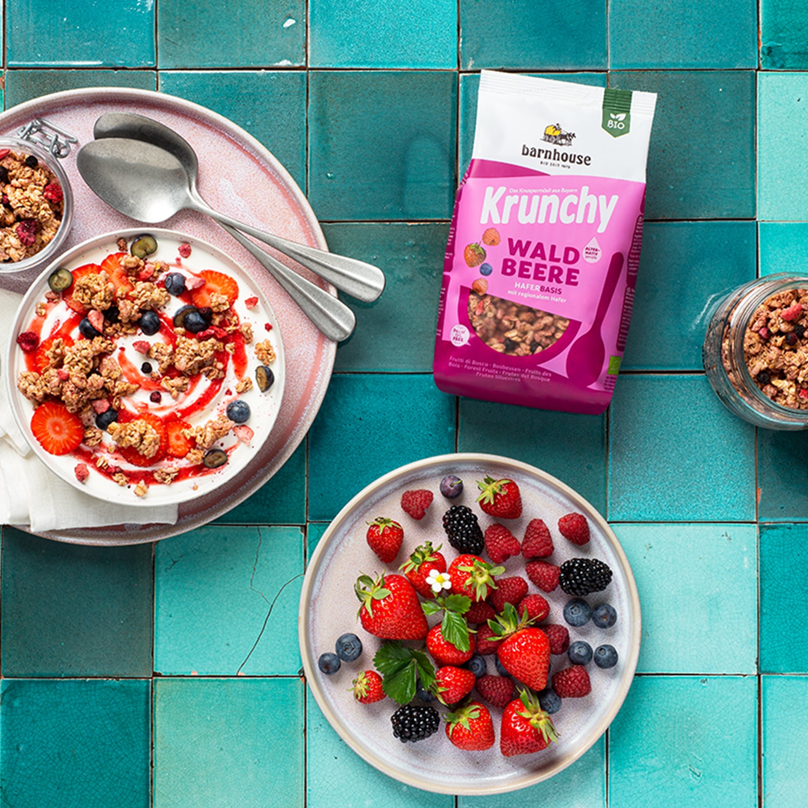 Blick von oben auf einen rosa-weißen Beutel Krunchy Waldbeere alternativ gesüßt von Bio-Knuspermüsli-Hersteller Barnhouse auf blauen Fliesen. Der Fokus liegt auf der Bowl, die mit dem Beeren-Müsli und Joghurt befüllt ist. Als Topping liegen Erdbeeren, Himbeeren und Heidelbeeren oben drauf, die mit einer Erdbeersoße betröpfelt sind. Links und rechts steht jeweils ein offenes Vorratsglas mit diesem Müsli und am unteren Bildrand ein Teller mit einem Mix aus Waldbeeren.