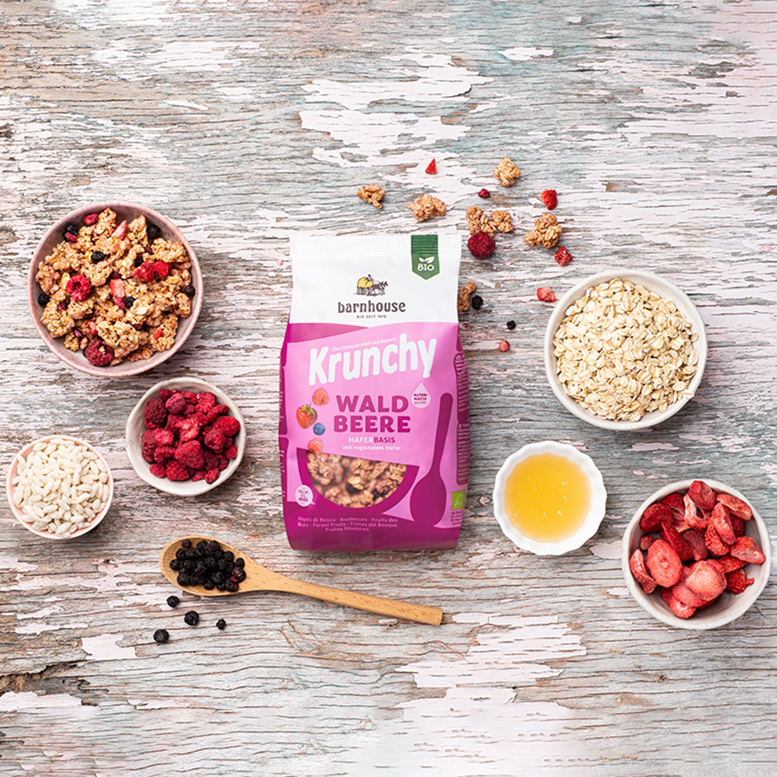 Blick von oben auf einen rosa-weißen Beutel Krunchy Waldbeere alternativ gesüßt von Bio-Knuspermüsli-Hersteller Barnhouse auf hellem Holztisch. Außenrum stehen fünf weiße Schalen mit Haferflocken, Sonnenblumenöl, Erdbeerscheiben, Himbeeren und Reispops. Eine weitere Schale ist mit dem Beeren-Knuspermüsli befüllt. Auf einem Holzlöffel liegen Johannisbeeren.