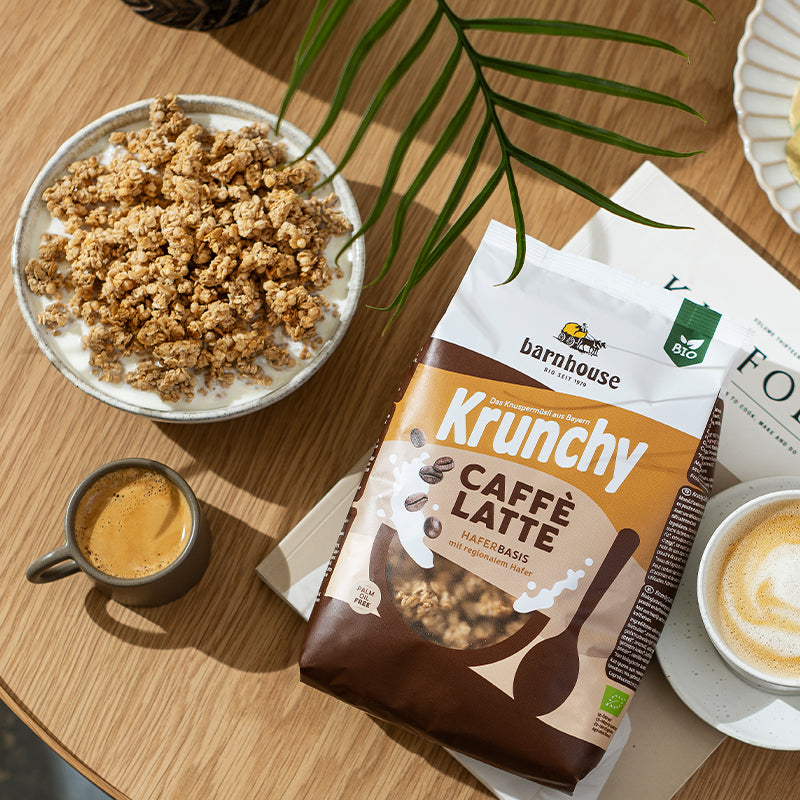 Ein Beutel Krunchy Caffè Latte von Bio-Knuspermüsli-Hersteller Barnhouse, der auf einer Zeitung liegt. Daneben befindet sich eine große, graue Schüssel mit Krunchy Caffè Latte sowie eine Tasse schwarzer Kaffee und ein Cappuccino mit Unterteller. Von oben blitzt ein dünnes, grünes Blatt einer Pflanze ins Bild. All die Elemente liegen auf einem runden Holztisch.