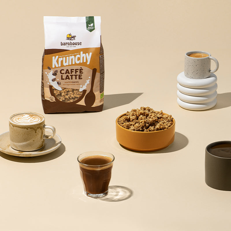 Beiger Untergrund, auf dem oben links ein Beutel Krunchy Caffè Latte von Bio-Knuspermüsli-Hersteller Barnhouse steht. In der Mitte steht eine orange-braune Schüssel mit diesem Müsli. Links eine gräuliche Tasse auf passendem Unterteller mit Cappuccino, dessen Milchschaum ein Blumenmuster zeigt. Vorne steht ein Kaffee mit Milch in einem durchsichtigen Glas und rechts ein schwarzer Kaffee in einer grauen Tasse. Oben rechts ist ein Milchkaffee in einer grauen, blau gepunkteten Tasse auf einem weißem Sockel.