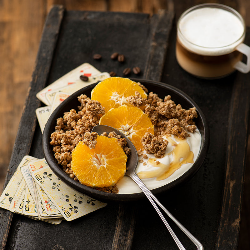 Schwarze Holzkiste auf einem braunen Holzboden. Darauf steht eine schwarze Schüssel mit Krunchy Caffè Latte von Bio-Knuspermüsli-Hersteller Barnhouse, Joghurt und Orangenscheiben. Neben der Müsli-Bowl liegen Kaffeebohnen, abgenutzte Pokerkarten und eine durchsichtige Tasse mit Milchkaffee.
