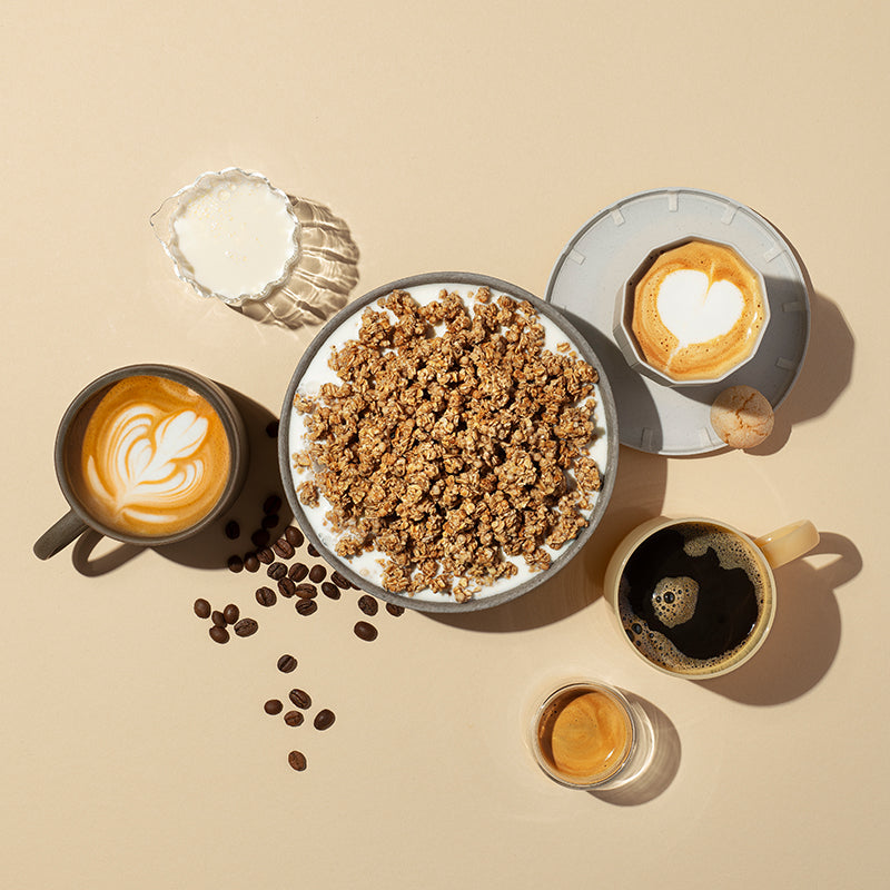 Einfarbiger, beiger Hintergrund, in dessen Fokus eine Schüssel mit Milch und Krunchy Caffè Latte von Bio-Knuspermüsli-Hersteller Barnhouse steht. Außenrum liegen einige Kaffeebohnen. Außerdem stehen dort noch ein Glaskaraffe mit Milch sowie ein Espresso und eine Tasse schwarzer Kaffee. Links steht ein Cappuccino mit Milchschaum in Blumenform in einer grauen Tasse. Auf der rechten Siete steht ein grauer Unterteller mit einem kleinen Keks und einer Cappuccino mit Milchschaum in Herzform.