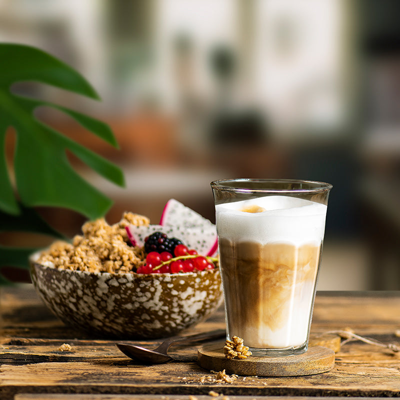 Ein Milchkaffee in einem durchsichtigen Glas auf einem hölzernen Untersetzer mit Müsli-Cluster, der auf einem Holztisch steht. Daneben liegt ein silberner Löffel und dahinter steht eine schön dekorierte Schüssel mit Krunchy Caffè Latte von Bio-Knuspermüsli-Hersteller Barnhouse - schön dekoriert mit Drachenfrüchten, Brombeeren und roten Johannisbeeren. Links blitzt noch eine großes, grünes Blatt einer Pflanze ins Bild.