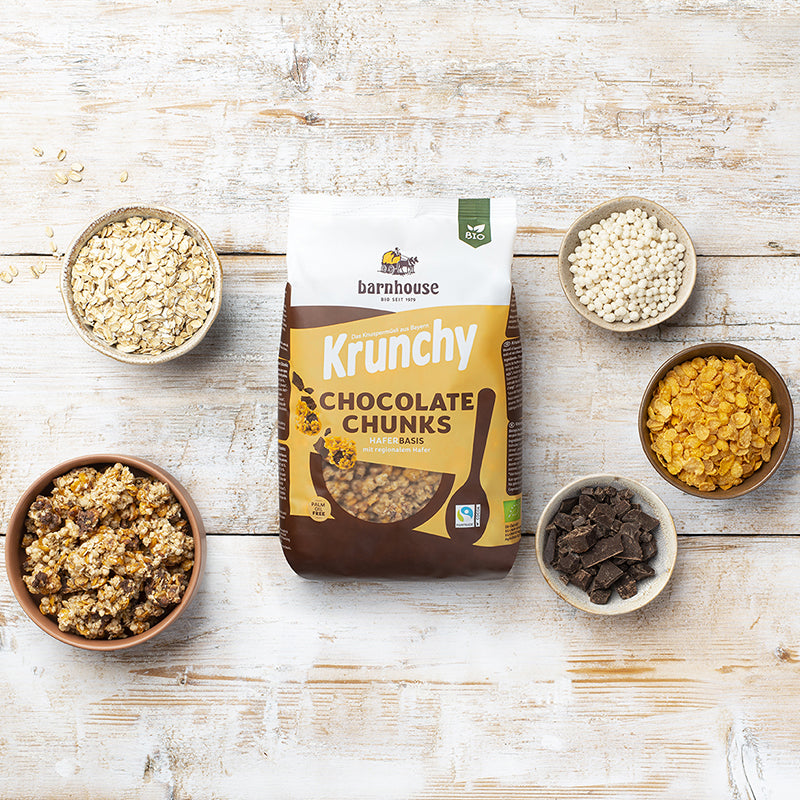 Beutel Krunchy Chocolate Chunks von Bio-Knuspermüsli-Hersteller Barnhouse auf einem weißen Holztisch. Links steht eine braune Schüssel mit diesem Müsli. Zudem stehen dort noch 3 weiße und eine braune Schale, die mit den Zutaten Haferflocken, Cornflakes, Schoko-Stückchen und Knusperreis gefüllt sind.