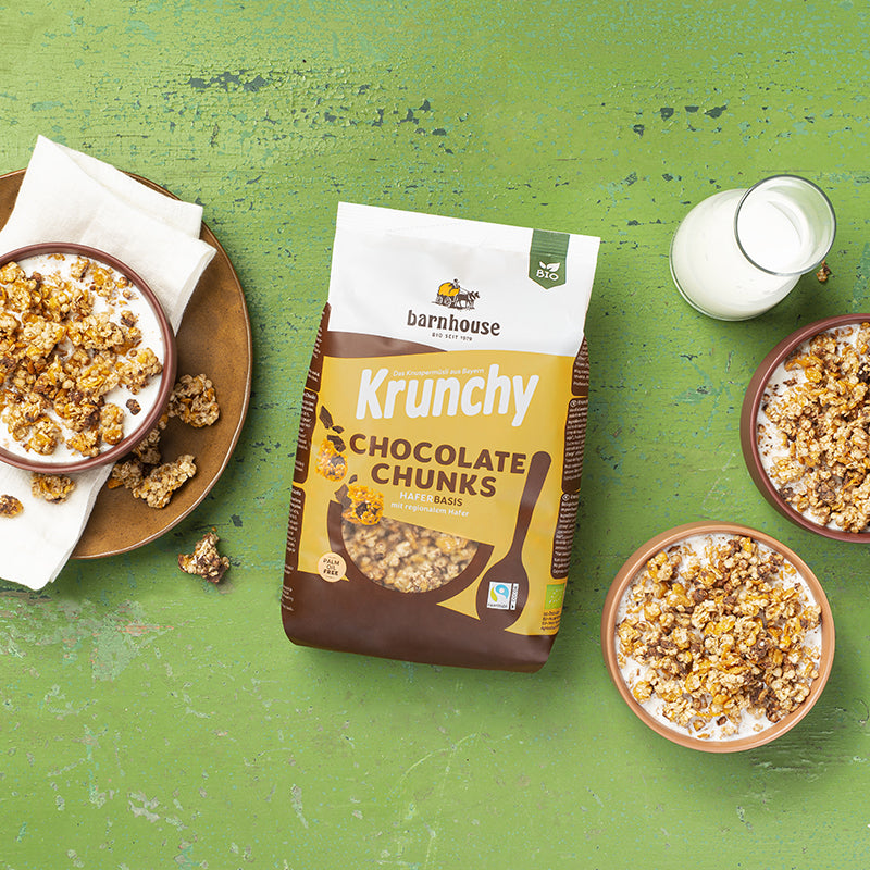 In der Mitte des grünen Holztisches, der schon etwas abblättert, liegt ein Beutel Krunchy Chocolate Chunks von Bio-Knuspermüsli-Hersteller Barnhouse. Rechts daneben steht eine Karaffe Milch sowie eine beige und eine braune Schüssel, befüllt mit diesem Müsli. Links ist ein brauner Unterteller zu sehen, auf dem eine weiße Serviette liegt. Auf dem Teller und daneben liegen ein paar Cluster. Auf der Serviette steht eine braune Schüssel, die mit diesem Müsli und Milch befüllt ist.