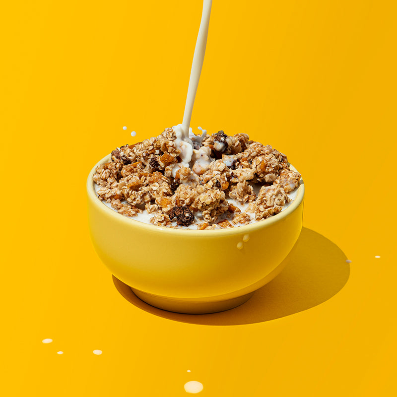 Gelbe Schüssel, die gut gefüllt mit Krunchy Chocolate Banana Chunks von Bio-Knuspermüsli-Hersteller Barnhouse ist. Vom oberen Bildrand wird ein Strahl Milch hinein geschüttet. Ansonsten sind noch ein paar Milchspritzer auf dem gelben Hintergrund zu sehen.