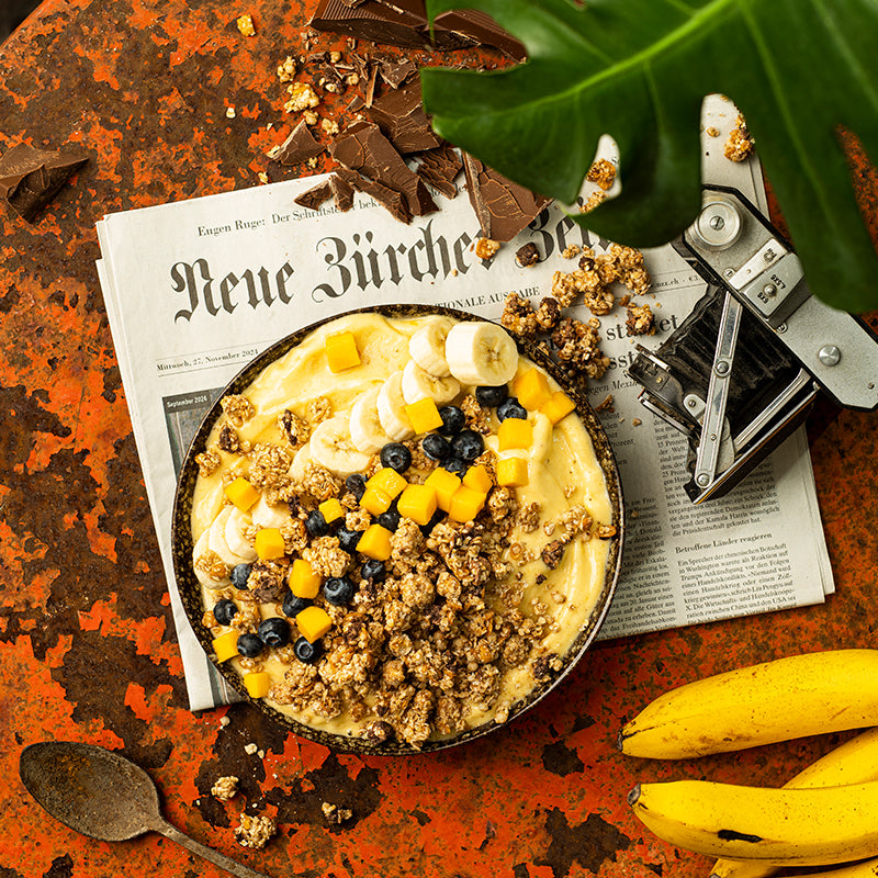 Eine Schüssel mit Krunchy Chocolate Banana Chunks von Bio-Knuspermüsli-Hersteller Barnhouse, Bananenscheiben, Ananasstücken, Heidelbeeren und Joghurt, die auf einer Zeitung liegt. Daneben liegen ein paar Cluster dieses Müslis sowie einzelne Schoko-Stückchen. Im Vordergrund liegen noch ein paar Bananen sowie ein Löffel auf dem rötlich-braunen Tisch.