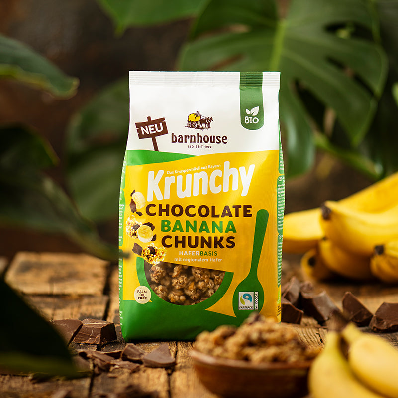 Blick von vorne auf einen Beutel Krunchy Chocolate Banana Chunks von Bio-Knuspermüsli-Hersteller Barnhouse, der auf einem braunen Holztisch steht. Im Hintergrund sind die großen Blätter einer grünen Pflanze zu sehen. Neben dem Müsli liegen einige Bananen und Schoko-Stückchen. Im Vordergrund steht noch eine unscharfe, braune Schüssel mit dem Müsli.