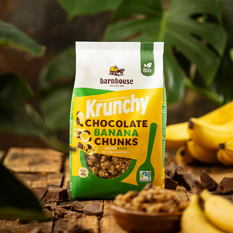 Blick von vorne auf einen Beutel Krunchy Chocolate Banana Chunks von Bio-Knuspermüsli-Hersteller Barnhouse, der auf einem braunen Holztisch steht. Im Hintergrund sind die großen Blätter einer grünen Pflanze zu sehen. Neben dem Müsli liegen einige Bananen und Schoko-Stückchen. Im Vordergrund steht noch eine unscharfe, braune Schüssel mit dem Müsli.