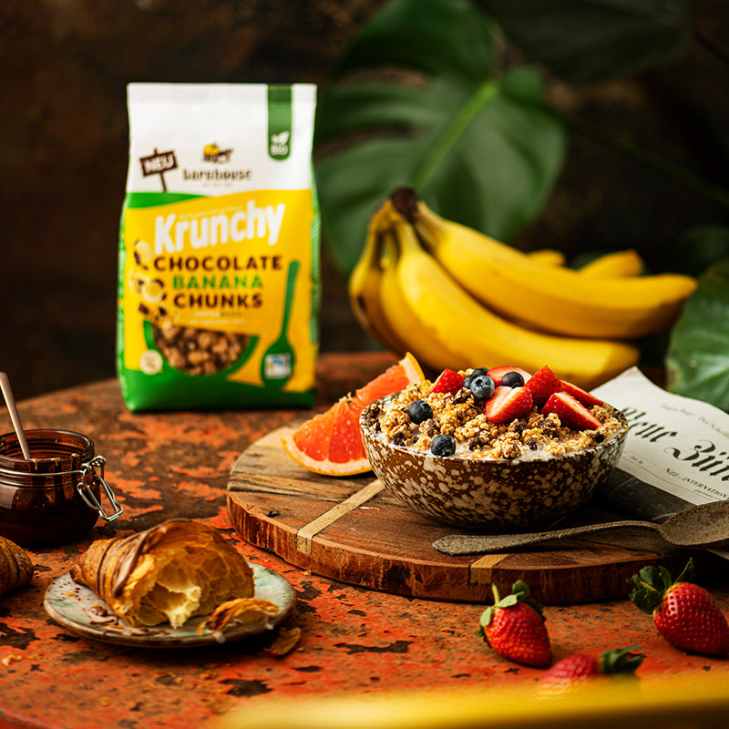 Bunt dekorierter, rötlicher Tisch mit einem Beutel Krunchy Chocolate Banana Chunks von Bio-Knuspermüsli-Hersteller Barnhouse. Außerdem auf dem Tisch: Bananen, ein Schoko-Croissant, Erdbeeren, eine Zeitung, eine Schüssel mit diesem Müsli sowie Erdbeeren und Heidelbeeren, eine Scheibe Blutorange. Im Hintergrund sind die Blätter einer grünen Pflanze zu sehen.