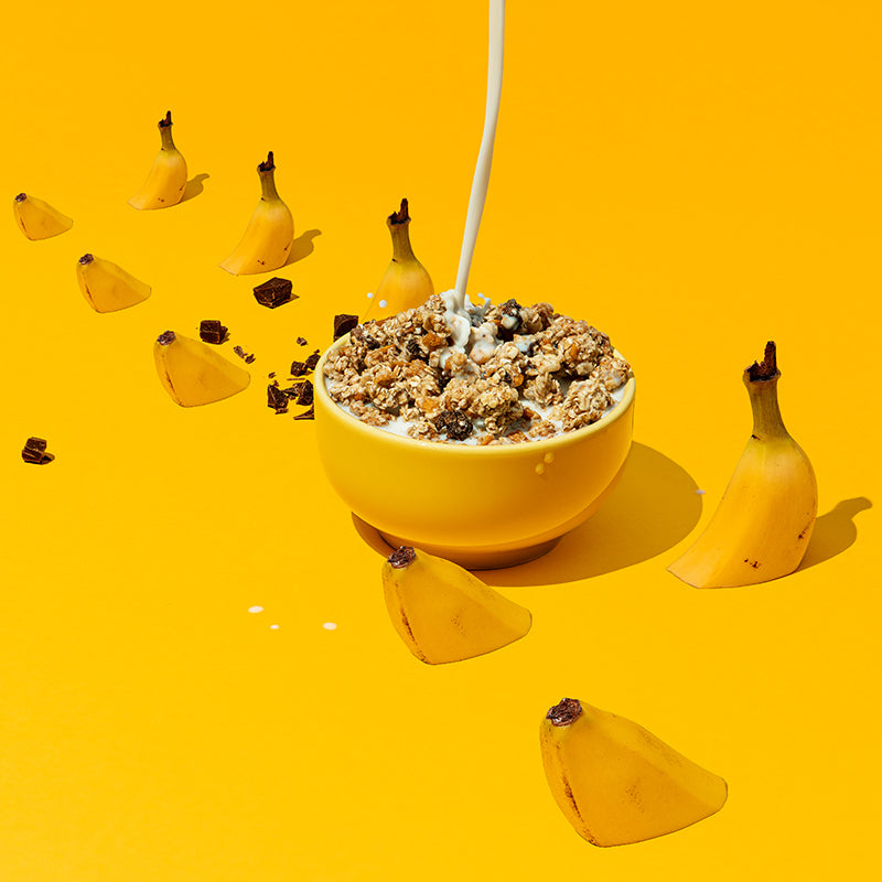 In der Mitte steht eine gelbe Schüssel mit Krunchy Chocolate Banana Chunks von Bio-Knuspermüsli-Hersteller Barnhouse. Von oben fließt ein Milchstrahl in die Bowl. Neben der Schüssel sind auch noch Schoko-Stückchen sowie angeschnittene Bananen zu sehen, die auf dem gelben Boden "sitzen".