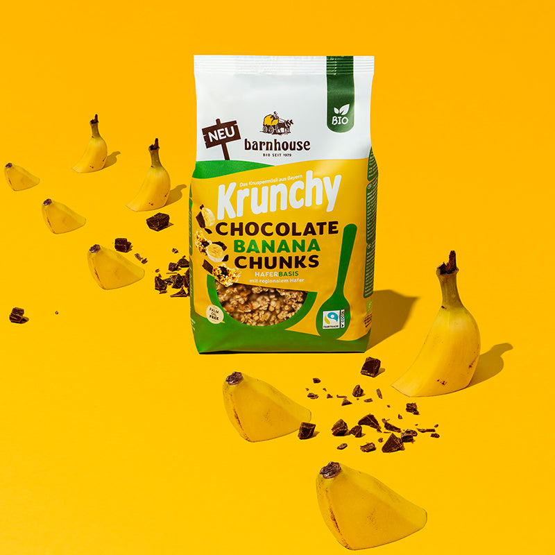 In der Mitte steht ein Beutel Krunchy Chocolate Banana Chunks von Bio-Knuspermüsli-Hersteller Barnhouse. Daneben sind Schoko-Stückchen sowie angeschnittene Bananen zu sehen, die auf dem gelben Boden "sitzen".