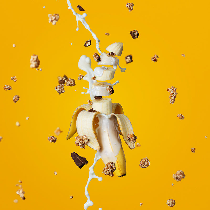 Eine zur Hälfte geschälte Banane, deren oberer Teil in Scheiben geschnitten ist. Von oben fließt ein Milchstrahl auf die Banane und läuft unten wieder aus dem Bild. Von oben fallen Knuspermüsli-Cluster, Bananen- und Schoko-Stücke - die Zutaten von Krunchy Chocolate Banana Chunks des Bio-Knuspermüsli-Herstellers Barnhouse - vor dem gelben Hintergrund nach unten.