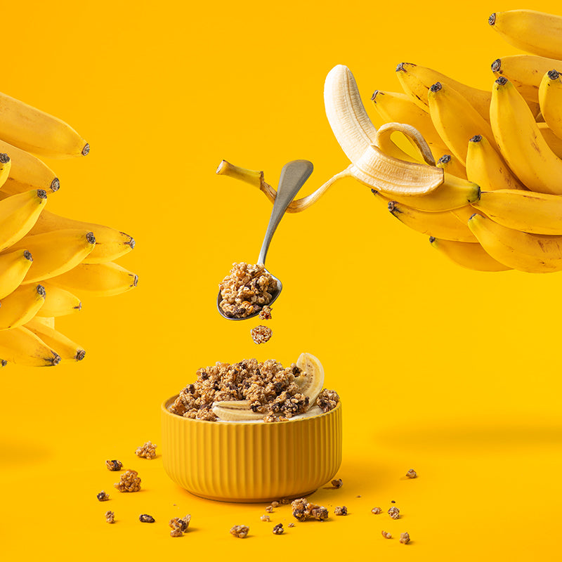 Eine gelbe, gerillte Schüssel, die mit Krunchy Chocolate Banana Chunks von Bio-Knuspermüsli-Hersteller Barnhouse und einer halben Banane befüllt ist. Daneben liegen ein paar Cluster dieses Müslis. Links und rechts ragen einige Bananen abgeschnitten in das Bild. Eine Banane ist ganz zu sehen und zur Hälfte geschält. Ein Teil der Schale wirkt wie ein Arm, der einen silbernen Löffel hält, von dem aus Müsli in die Schüssel fällt.