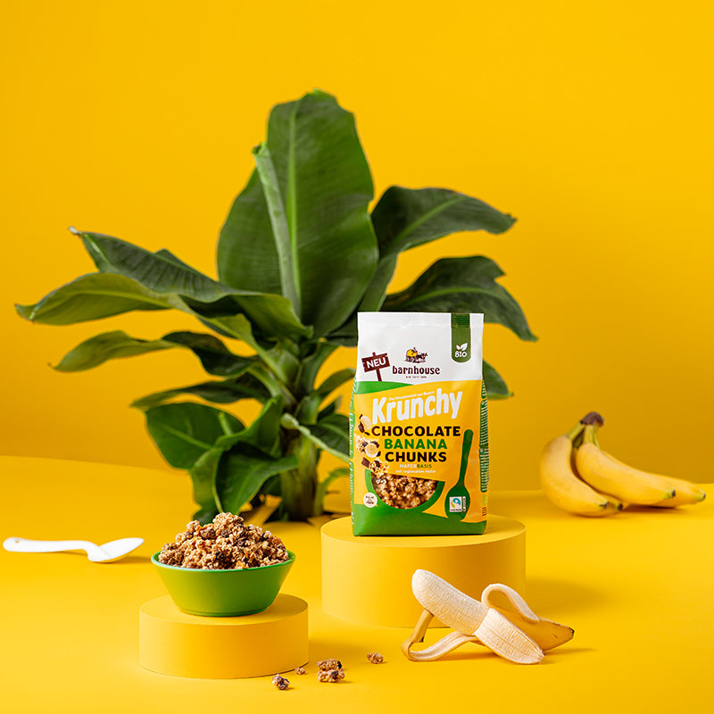 Gelber Hintergrund mit einer grünen Pflanze. Rechts daneben liegen ein paar Bananen und links ein weißer Löffel. In der Bildmitte stehen ein Beutel Krunchy Chocolate Banana Chunks von Bio-Knuspermüsli-Hersteller Barnhouse und eine grüne Schüssel mit diesem Müsli auf zwei verschiedenen gelben Sockeln. Vorne liegen noch ein paar Cluster sowie eine "Chiller"-Banane, die zur Hälfte geschält ist und einen Teil ihrer Schale als Arm nutzt, auf dem sich die restliche Banane lässig abstützt.