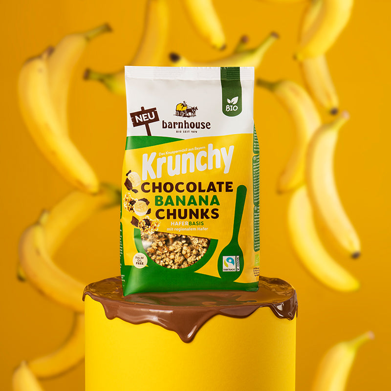 Beutel Krunchy Chocolate Banana Chunks von Bio-Knuspermüsli-Hersteller Barnhouse, der auf einem gelben Sockel steht, dessen Oberfläche mit geschmälzter Schokolade überdeckt ist. Zwischen dem Beutel und dem gelben Hintergrund fallen einige ungeschälte Bananen vom Himmel.