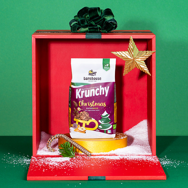Ein rote, geöffnete Geschenkbox mit goldenem Stern, etwas künstlichem Schnee, einer Zuckerstange, einem Tannenzweig und einer grüner Schleife. Im Geschenk steht ein Beutel Krunchy Christmas von Bio-Knuspermüsli-Hersteller Barnhouse auf einem goldenen Sockel. Die Geschenkbox steht vor einem grünen Hintergrund.