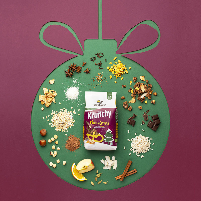 Violetter Hintergrund, in dem die Form einer Christbaumkugel durch grüne Farbe eingezeichnet ist. In diesem Innenraum befindet sich ein Beutel Krunchy Christmas von Bio-Knuspermüsli-Hersteller Barnhouse sowie die einzelnen Zutaten dieses Müslis - Apfelstücke, Orangenscheiben, Haferflocken, Kokoschips, dunkle Schoko-Stückchen Und Co.