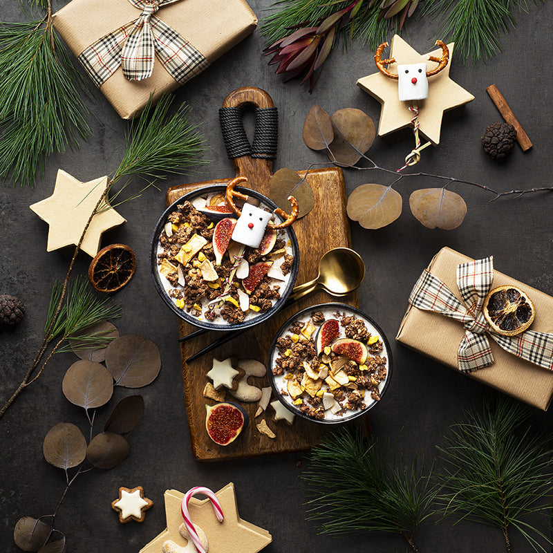 Schwarzer Tisch, der mit Geschenken, Tannenzweigen, Marshmallows, Orangenscheiben, und Co. sehr weihnachtlich geschmückt. In der Mitte liegt ein braunes Holzbrett, auf dem sich 2 Schüssel befinden, die mit Krunchy Christmas von Bio-Knuspermüsli-Hersteller Barnhouse befüllt sind. Verfeinert sind die Bowls mit Feigen und Marshmallows mit Rentiergesicht. Außerdem liegen noch ein Zimtstern, Vanillekipferl, eine halbe Feige sowie 2 goldene Löffel auf dem Brett. Ein wirklich weihnachtlicher Anblick.