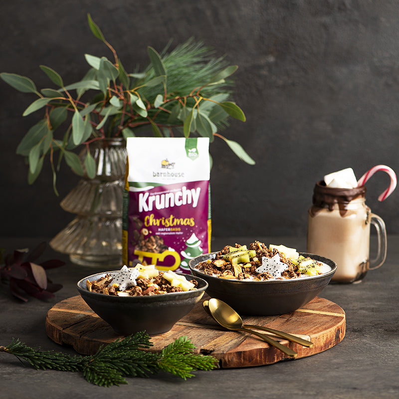 Weihnachtlich dekoriertes Bild, bei dem der Beutel Krunchy Christmas von Bio-Knuspermüsli-Hersteller Barnhouse im Fokus steht - mit grauem Hintergrund. Auf der grauen Tischplatte liegt eine braune Holzplatte mit 2 goldenen Löffeln & 2 schön dekorierten, grauen Müslischüsseln. Neben unserem Weihnachts-Müsli sind dort verschiedene Früchte in Sternform mit drin. Außerdem liegt noch ein Tannenzweig auf dem Tisch. Im Hintergrund stehen noch eine durchsichtige Vase mit grünen Blättern sowie ein Schoko-Milchshake.