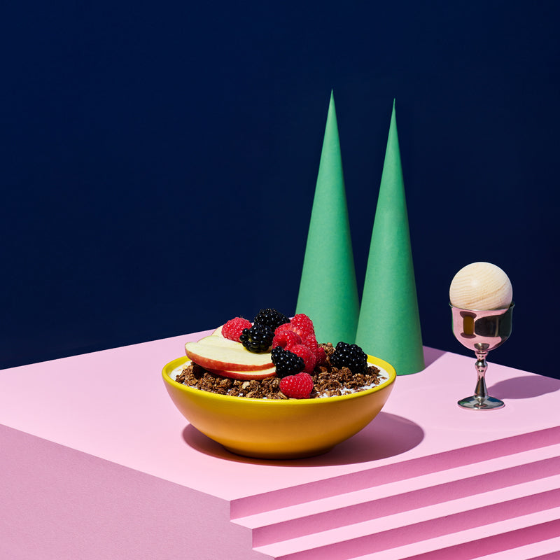 Rosa Treppe, auf der eine gelbe Schüssel mit Krunchy Cocoa weniger Zucker, Milch, Himbeeren, Brombeeren und Apfelscheiben steht. Daneben ein silberner Eierbecher mit Holzkugel sowie 2 dunkelgrüne, hohe Kegel.