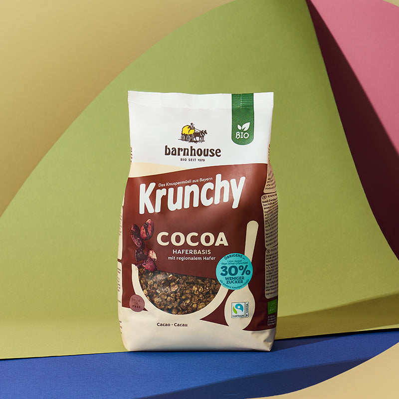 Braun-beiger Beutel Krunchy Cocoa weniger Zucker von Bio-Knuspermüsli Hersteller Barnhouse vor einem bunten Hintergrund mit einer gelben, grünen, roten, blauen und brauen Fläche.