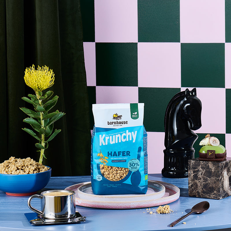 Blauer Beutel Krunchy Hafer weniger Zucker von Barnhouse in in zwei bunt gesprenkelten Tellern auf einem braunen Holztisch. Links daneben eine blaue Schüssel mit Milch und dem besagten Bio-Knuspermüsli, dahinter eine gelbe Blume . Zudem sind noch eine gefüllte, silberne Espressotasse, eine Pferdekopf Schach-Skulptur, ein Löffel und eine Deko-Skulptur. Im Hintergrund eine Wand mit dunkelgrün-gräulichem Schachmuster sowie einem dunkelgrünen Vorhang.