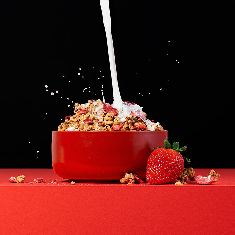 Rote Schüssel - gut gefüllt mit Krunchy High Protein von Barnhouse - in die ein kräftiger Strahl Milch geschüttet wird. Neben der Schüssel liegt eine Erdbeere sowie Cluster und Erdbeerscheiben vom erwähnten Bio-Knuspermüsli.