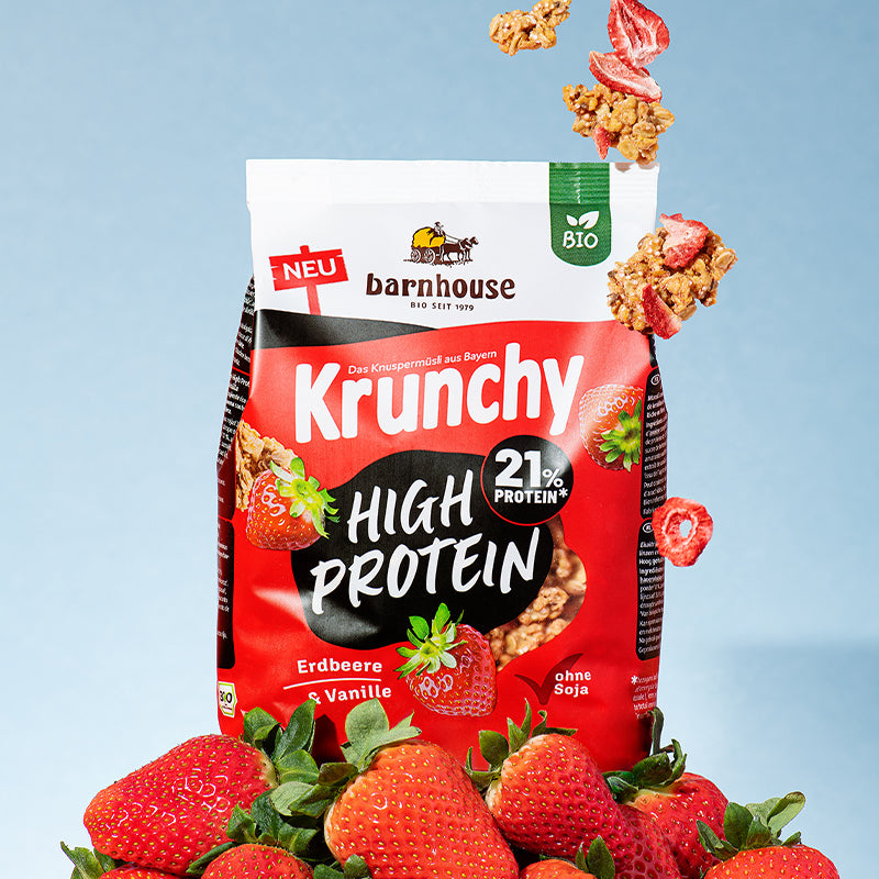 Beutel Krunchy High Protein von Bio-Knuspermüsli-Hersteller Barnhouse, der auf einem Berg aus Erdbeeren steht. Von oben fallen Knuspermüsli-Cluster und Erdbeerscheiben nach unten.