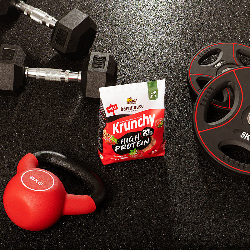Beutel Krunchy High Protein von Bio-Knuspermüsli-Hersteller Barnhouse, der in einem Fitnessraum zwischen einer 8kg Kugelhantel sowie 2 Hanteln und 2 Hantelscheiben steht.