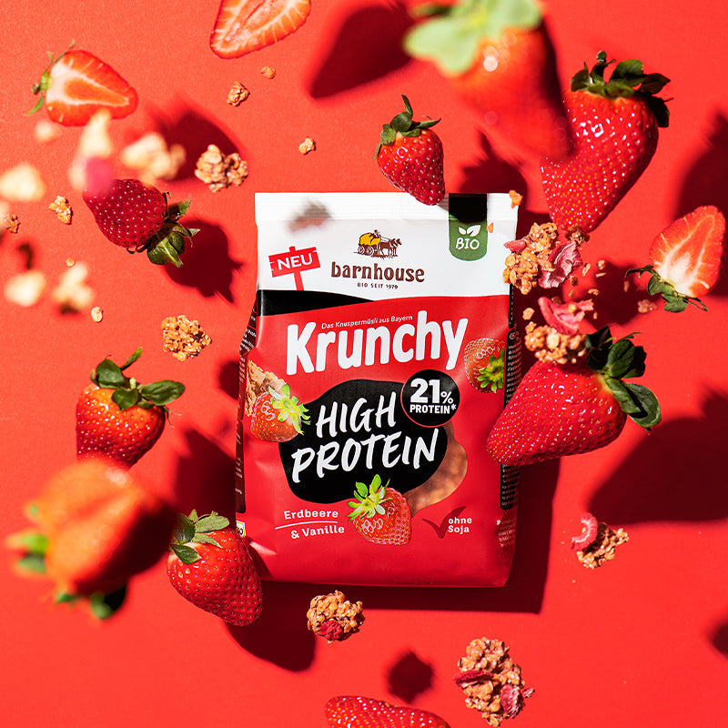 Beutel Krunchy High Protein von Barnhouse, der auf einer roten Fläche liegt. Von oben fallen jede Menge Erdbeeren (ganz und geteilt) sowie Knuspermüsli-Cluster mit Erdbeerscheiben auf den Beutel.