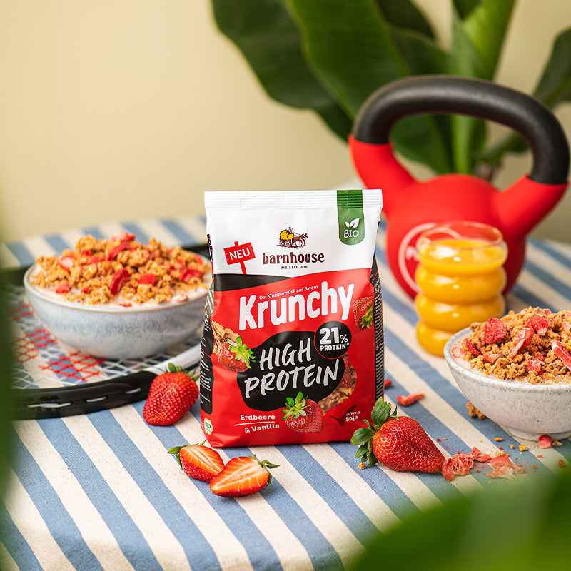 Ein Frühstückstisch mit blau-weißer Decke. In der Mitte steht ein Beutel Krunchy High Protein von Barnhouse. Außenrum sind Erdbeeren (ganz, geteilt und Scheiben). ein Glas mit Orangensaft sowie eine Kugelhantel zu sehen. Außerdem liegt links noch ein Tennisschläger auf dem Tisch, auf dem eine graue Schüssel mit besagtem Bio-Knuspermüsli steht. Auf der rechten Seite steht nochmal eine mit Krunchy High Protein befüllte Schüssel, während vorne, hinten und seitlich grüne Pflanzen zu sehen sind.
