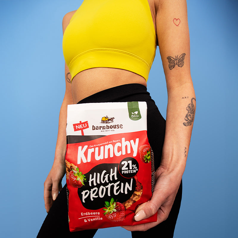 Körper einer jungen, durchtrainierten und tätowierten Frau im Sport-Outfit, der einen Beutel Krunchy High Protein von Bio-Knuspermüsli-Hersteller Barnhouse in der linken Hand hält.