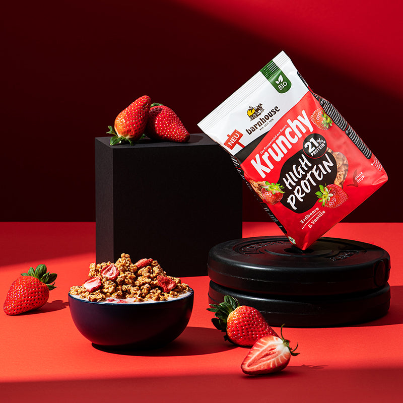Beutel Krunchy High Protein von Barnhouse, der im 45 Grad Winkel (nach links geneigt) auf 2 Hantelgewichten steht. Links daneben befindet sich ein schwarzes Achteck, aus dem 2 Erdbeeren liegen. Auf dem roten Untergrund befinden sich noch 2 weitere, ganze Erdbeeren sowie ein Erdbeer-Hälfte. Links vorne steht eine dunkle Schale mit dem besagten Bio-Knuspermüsli (Cluster + Erdbeerscheiben) und Milch.