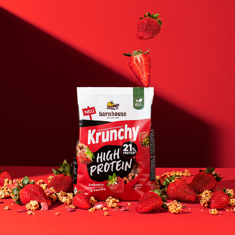 Blick von vorne auf einen Beutel Krunchy High Protein von Barnhouse, der auf auf einer roten Fläche steht. Von oben fallen 2 Erdbeeren (eine ganze und eine Hälfte) herunter und außenrum liegen jede Menge Erdbeeren sowie Cluster vom erwähnten Bio-Knuspermüsli.