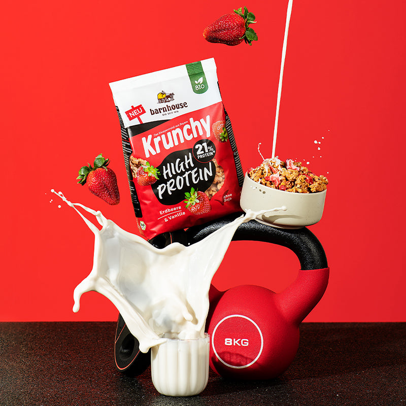 Blick von vorne auf einen Beutel Krunchy High Protein von Bio-Knuspermüsli-Hersteller Barnhouse, der auf einem Hantelgewicht und einer Kugelhantel steht. Davor steht ein Glas, aus dem Milch herausspritzt. Außerdem befindet sich noch eine Schüssel mit Krunchy High Protein auf der Kugelhantel, in die von oben ein Strahl Milch fließt. Zudem fliegen noch 2 Erdbeeren durch das Bild.