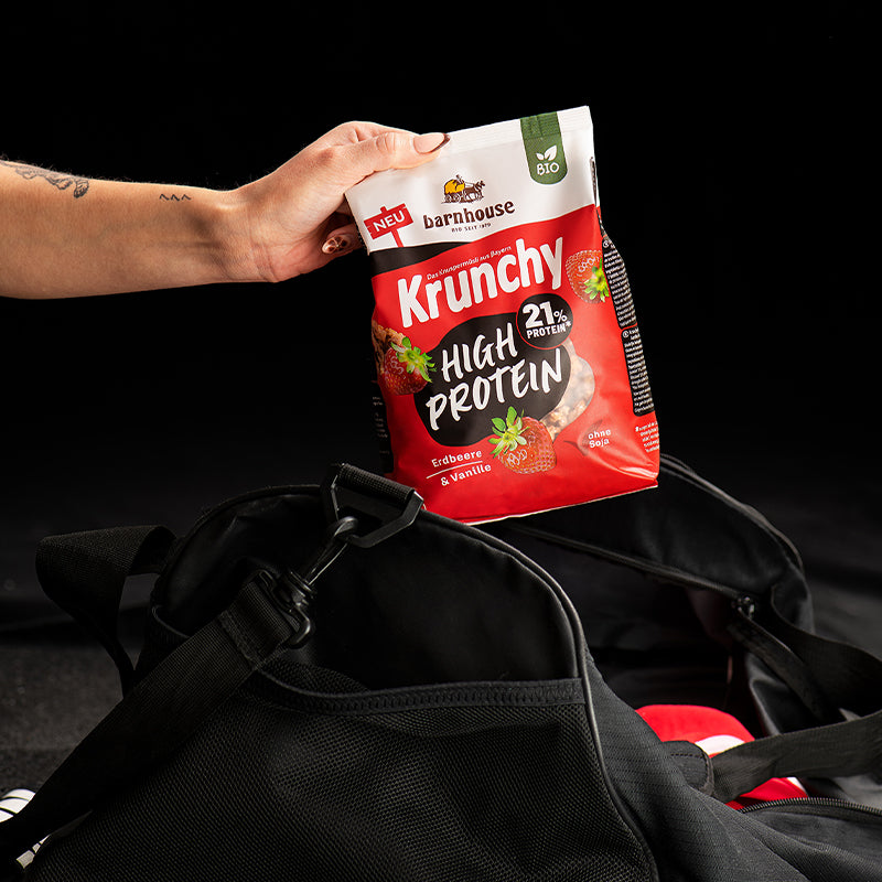 Arm einer tätowierten Frau mit gemachten Fingernägeln, die einen Beutel Krunchy High Protein von Bio-Knuspermüsli-Hersteller Barnhouse aus ihrer schwarzen Sporttasche holt.