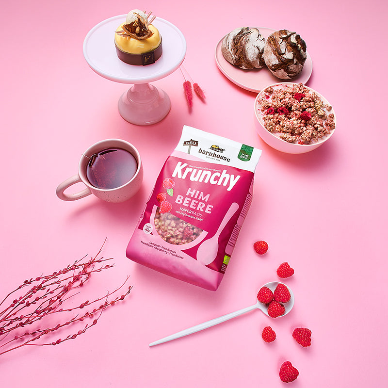 Blick von oben auf den beerenfarbigen, rosa-weißer Beutel Krunchy Himbeere von Bio-Knuspermüsli-Hersteller Barnhouse, der auf einem rosafarbenen Tisch liegt. Außenrum stehen eine Schale mit diesem Müsli und Milch, eine Tasse mit Früchtetee, ein Teller mit 2 Leib Brot, sowie ein Eierlikör-Dessert auf einem weißen Teller und ein weißer Löffel mit Himbeeren.