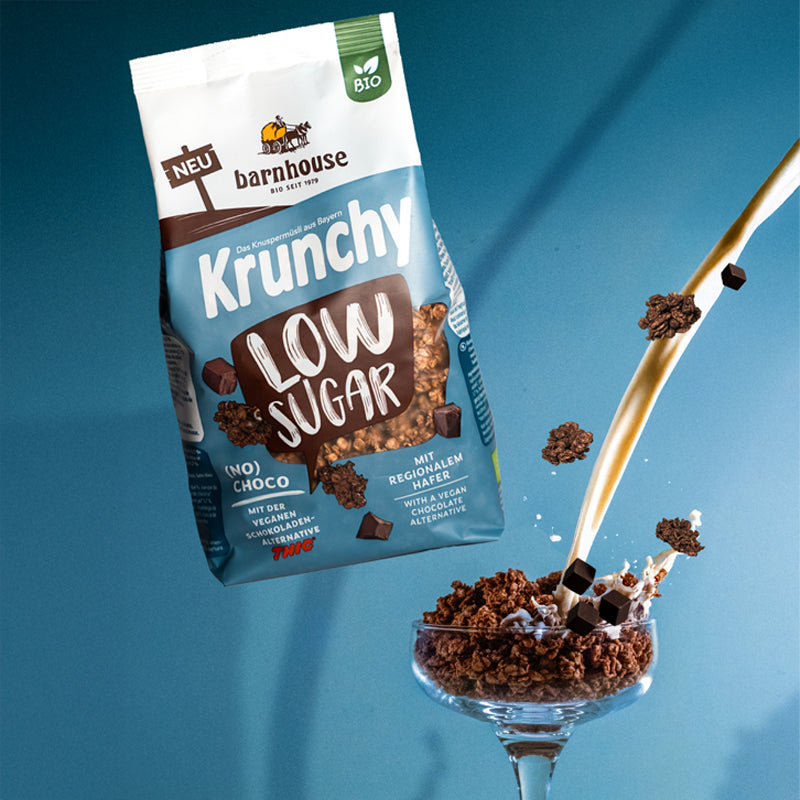 Beutel Krunchy Low Sugar (No) Choco, der über einem Glas schwebt, das mit Knuspermüsli-Clustern befüllt ist. Vom rechten Bildrand fließt ein Milchstrahl ins Glas und "Schoko"-Cluster fliegen herum.