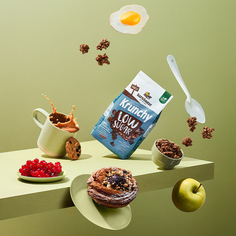 Blick voller Schwung von vorne auf ein grünes Holzbrett. Alle Elemente auf dem Bild fliegen durch die Luft: Braun-blau-weißer Beutel Krunchy Low Sugar (No) Choco von Bio-Knuspermüsli-Hersteller Barnhouse, Schale mit diesem Müsli, Krunchy-Cluster, weißer Löffel, Spiegelei, Tasse mit Kaffee, Schoko-Keks, Teller mit Johannisbeeren, Teller mit Nussschnecke und ein grüner Apfel.