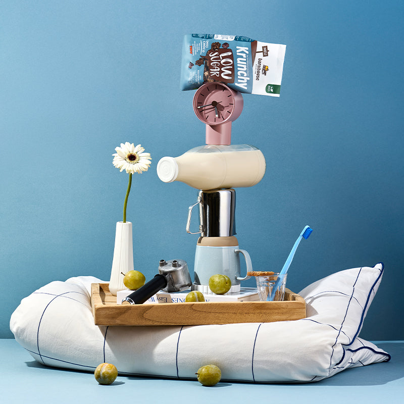 Blick von vorne auf ein Holzbrett, das auf einem blau-weiß karierten Kissen steht. Auf dem blauem Boden liegen 2 Birnen. Auf dem Holzbrett stehen ein Glas mit blauer Zahnbürste, eine weiße Vase mit weißer Blume und ein Kaffeesieb. Daneben ein Turm aus einem Buch, einer Tasse, einer Milchkanne, einer Milchflasche sowie einer Uhr und ganz oben liegt ein Beutel Krunchy Low Sugar (No) Choco von Bio-Knuspermüsli-Hersteller Barnhouse.