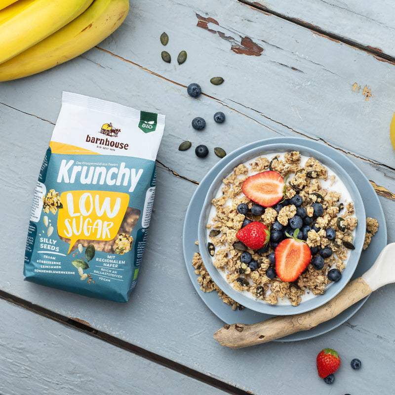 Blick von oben auf einen gelb-weiß-blauen Beutel Krunchy Low Sugar Silly Seeds von Bio-Knuspermüsli-Hersteller Barnhouse. Daneben steht eine Müslischale, die mit Milch und dem zuckerarmen Müsli befüllt ist. Frische Erdbeerscheiben und Heidelbeeren sorgen für eine schöne Farbkombi. Auf dem Unterteller liegt außerdem noch ein Holzlöffel sowie Müsli-Cluster. Auf dem blau gestrichenen Holztisch findet man noch Erdbeeren, Heidelbeeren, Bananen und Kürbiskerne verteilt.