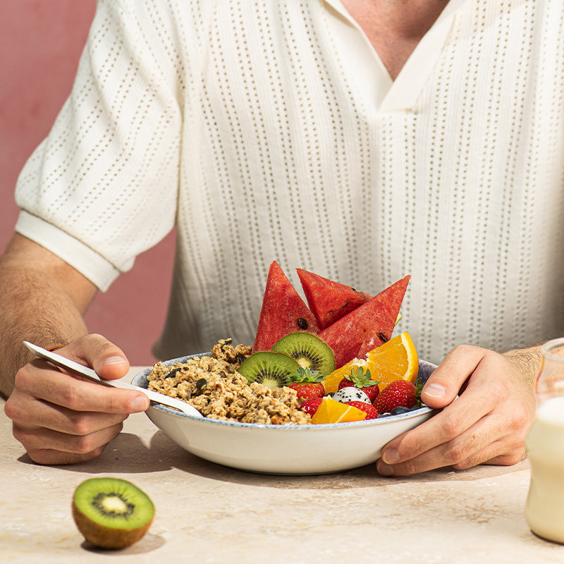 Eine sehr exotisch angerichtete Müsli-Bowl mit Krunchy Low Sugar Silly Seeds von Bio-Knuspermüsli-Hersteller Barnhouse - mit Kiwi, Wassermelone, Orange, frischen Erdbeeren, Sternenfrucht & Heidelbeeren als Topping. Im Hintergrund sitzt ein Mann am weißen Marmortisch, der mit der linken Hand die Schüssel festhält & mit der rechten Hand einen weißen Löffel in die Schüssel hält. Die Person ist nur von der Hüfte bis zum Hals sichtbar. Vorne links liegt eine halbe Kiwi & rechts ist ein Glas Milch angeschnitten.